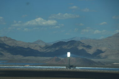 A farm of solar panels with a solar panel tower reflecting light is located on flat ground with rocky hills behind them.